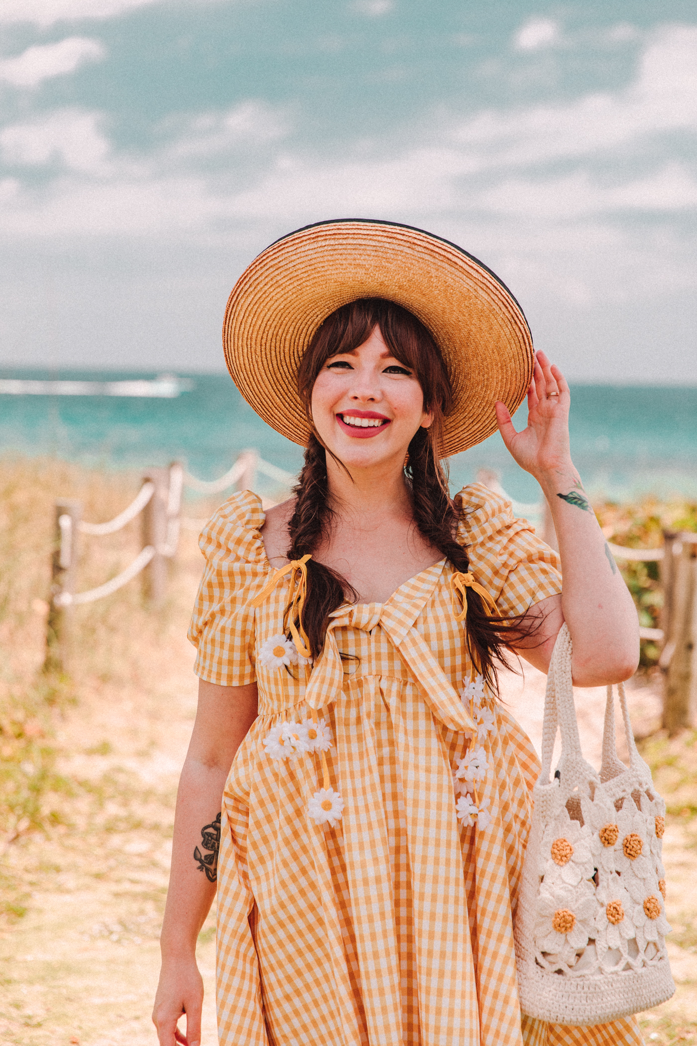 woman wearing yellow gingham dress, hart, flats, and holding a crochet bag at the beach