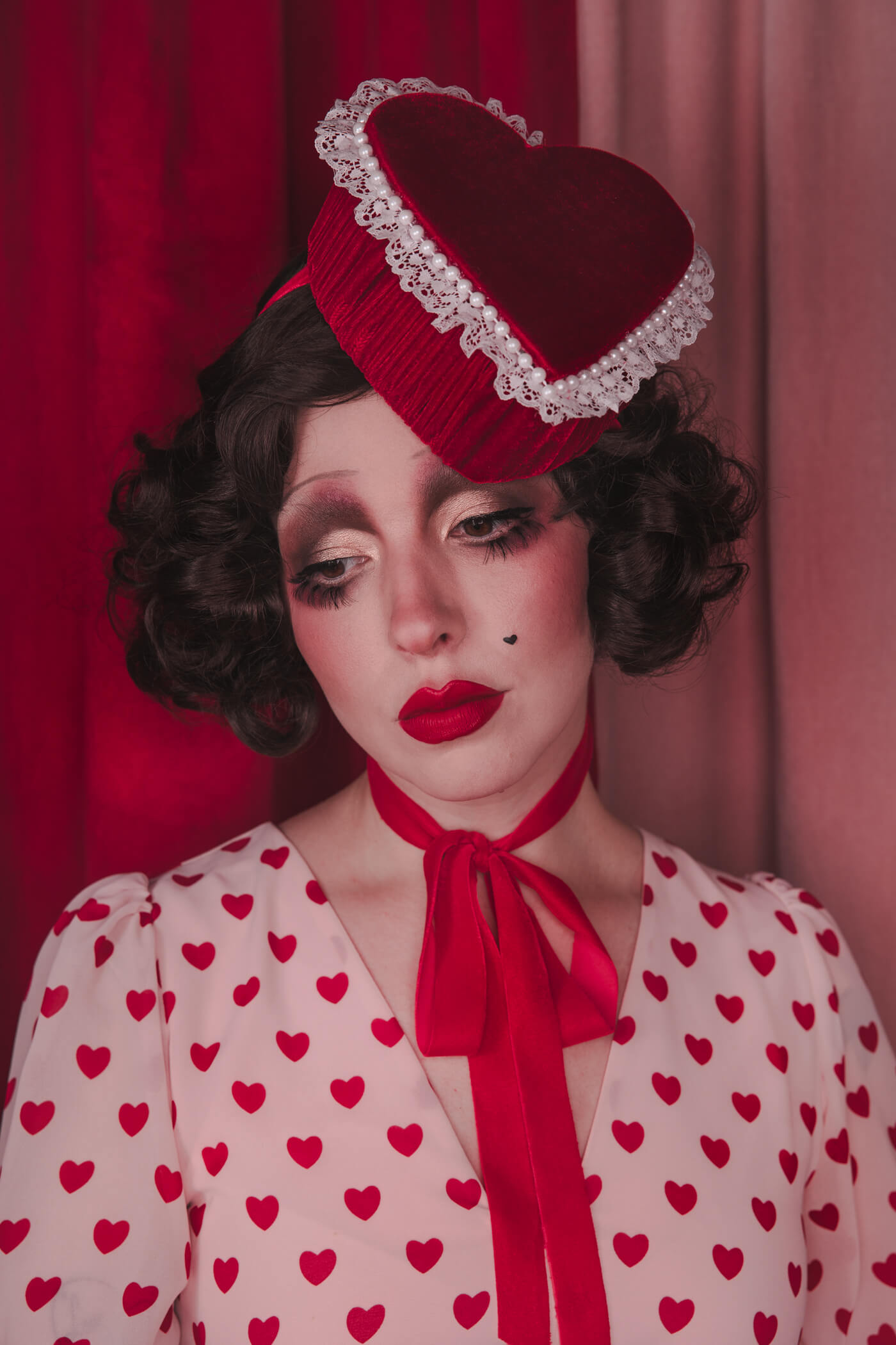 Pale girl with dark 20s 30s style hair and makeup, wearing a red velvet heart shaped box hat