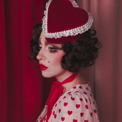 Pale girl with dark 20s 30s style hair and makeup, wearing a red velvet heart shaped box hat