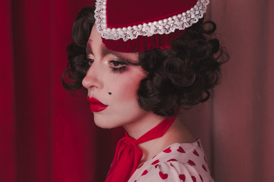 Pale girl with dark 20s 30s style hair and makeup, wearing a red velvet heart shaped box hat