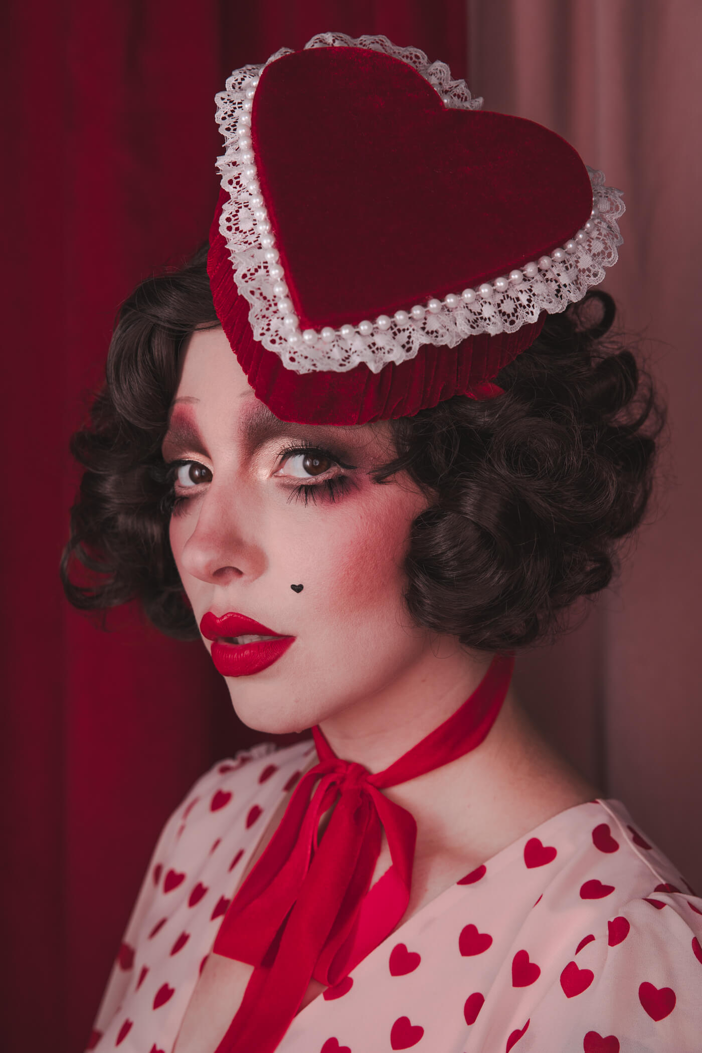 Pale girl with dark 20s 30s style hair and makeup, wearing a red velvet heart shaped box hat